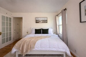 A bedroom with a bed, a bedside table, a lamp, a window with curtains, a door with frosted glass panels, and a painting on the wall. - Family-Friendly 4BR Game Room & Near Downtown (Idaho Falls)