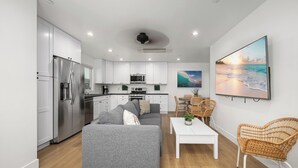 Cocina moderna con armarios blancos, refrigerador de acero inoxidable y comedor con sillas de mimbre y una mesa blanca.