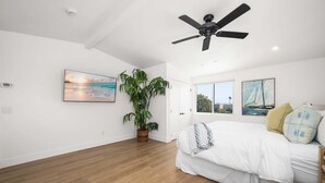 Un dormitorio con una cama, ventilador de techo, ventana grande y una planta en maceta.