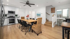 A modern kitchen with white cabinets, a microwave, and a stove. There is a dining area with a wooden table and wicker chairs. A ceiling fan is visible, and a staircase leads to another level. - Large Newport Beach Home Walk to the Beach (Newport Beach)