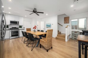A modern kitchen with white cabinets, a microwave, and a stove. There is a dining area with a wooden table and wicker chairs. A ceiling fan is visible, and a staircase leads to another level. - Large Newport Beach Home Walk to the Beach (Newport Beach)