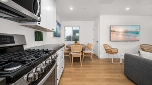 A modern kitchen with a stove, microwave, and white cabinets. There is a dining area with a table and chairs, and a gray sofa in the living room. - Large Beachside Escape in Prime Location (Newport Beach)