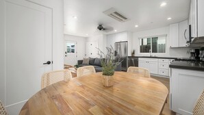 A modern kitchen with white cabinets, a stainless steel refrigerator, and a dining area with a wooden table and wicker chairs. - Large Beachside Escape in Prime Location (Newport Beach)