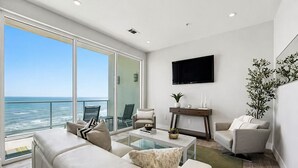 A modern living room with a large glass door leading to a balcony overlooking the ocean, a flat-screen TV mounted on the wall, and a minimalist decor style. - Emerald Suite 607 ~ Luxury 3BR Oceanfront + Full Resort Amenities Included (Galveston)