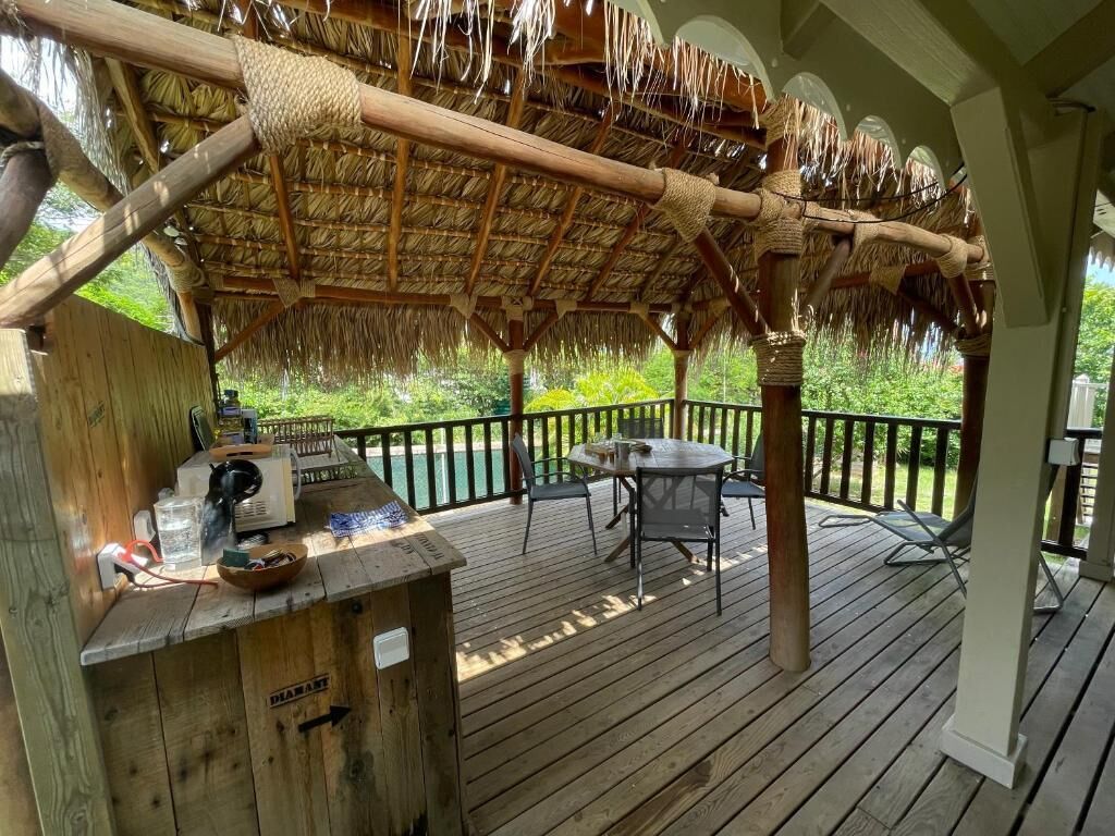 A wooden deck with a thatched roof, a table with chairs, and a small wooden structure with a sign.