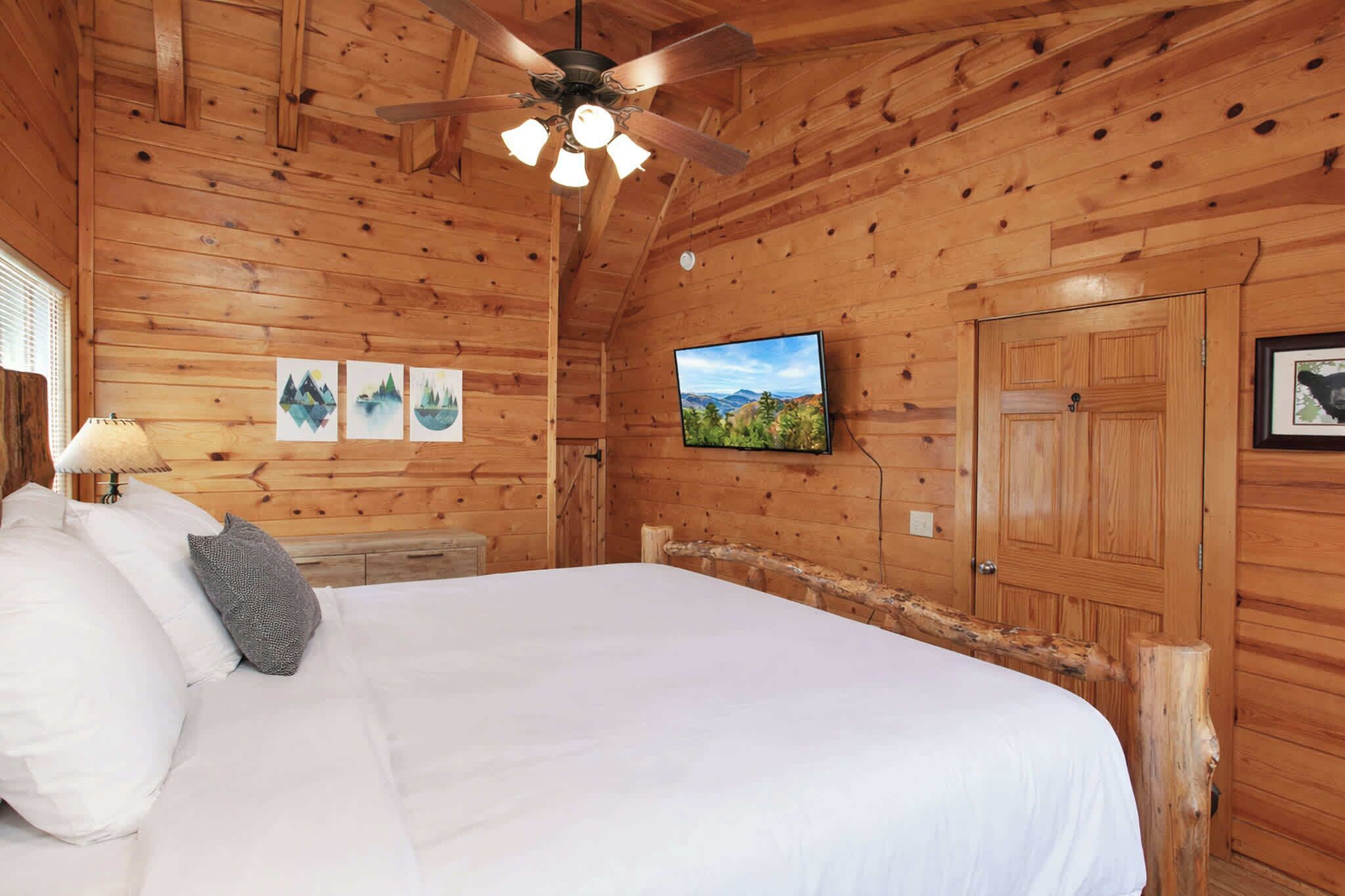 A wooden cabin bedroom with a bed, a ceiling fan, a TV, and a door.