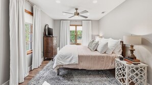 Une chambre à coucher comprenant un lit, un ventilateur de plafond, une commode, une table de chevet et une fenêtre avec des rideaux.