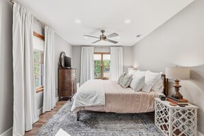 Un dormitorio con una cama, ventilador de techo, cómoda, mesita de noche y ventana con cortinas.