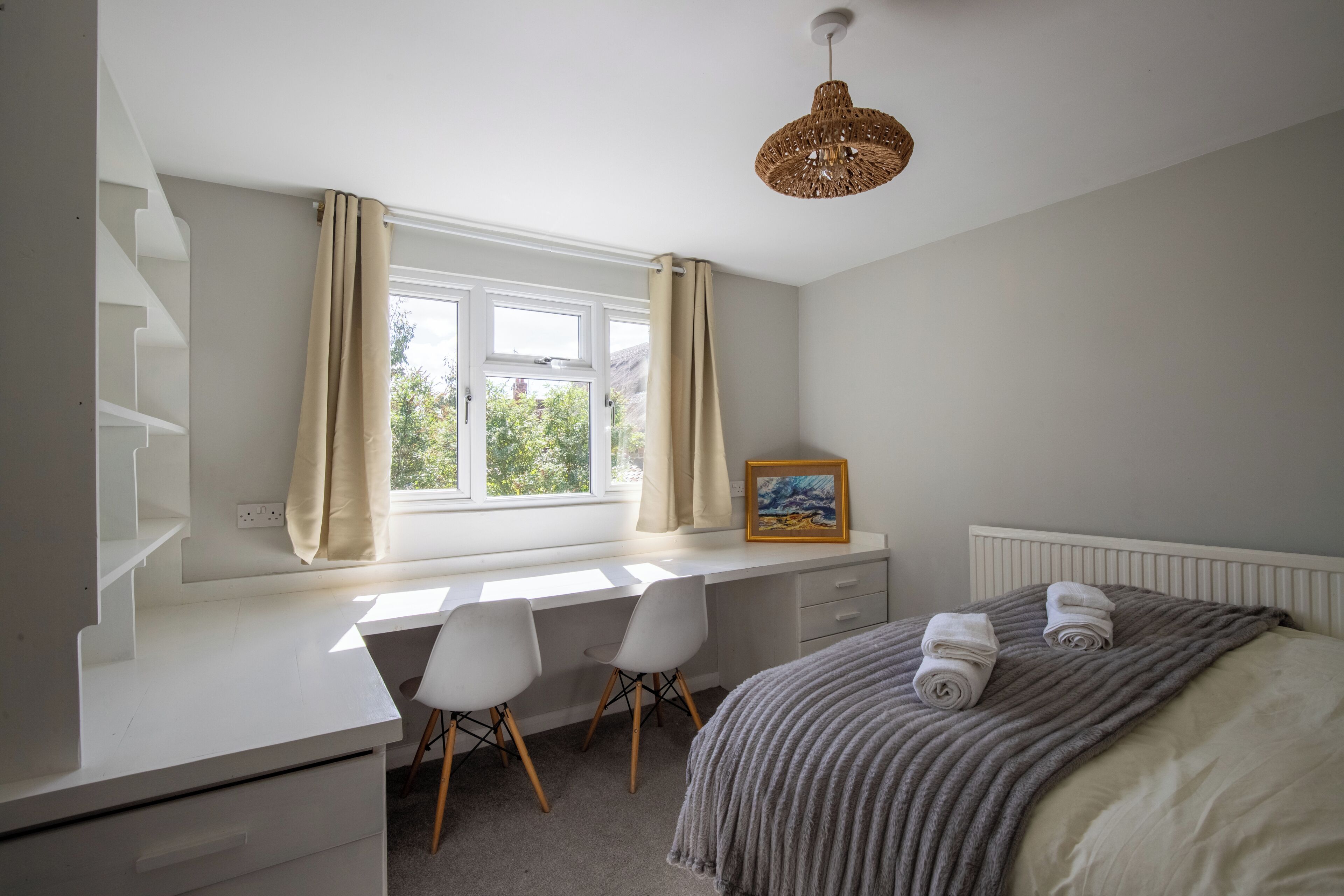 Une chambre à coucher avec un lit, un bureau avec deux chaises, une fenêtre avec des rideaux, un cadre photo et un luminaire suspendu.