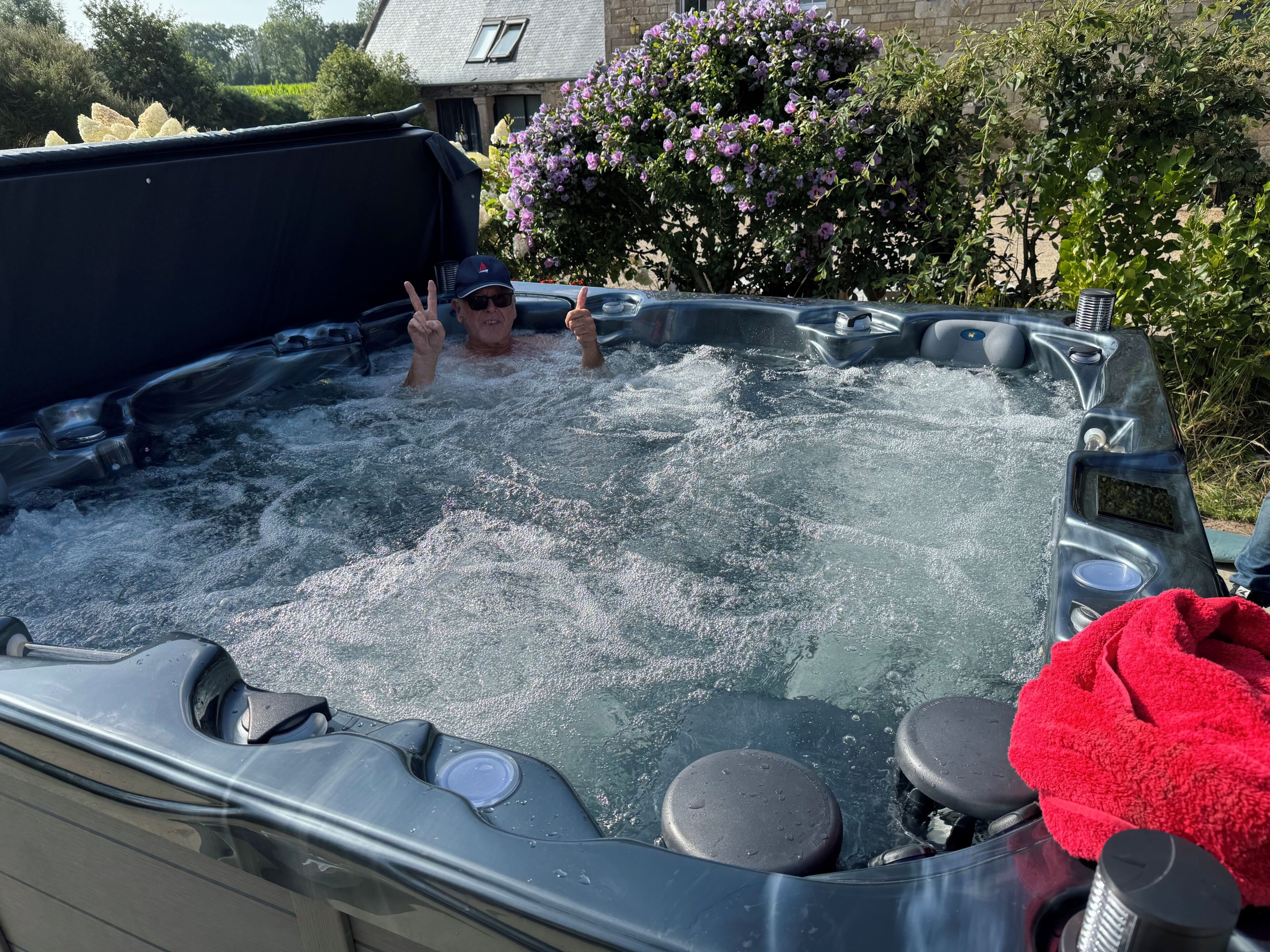 Un bain à remous avec une personne à l’intérieur, entouré de verdure et avec une maison en arrière-plan.