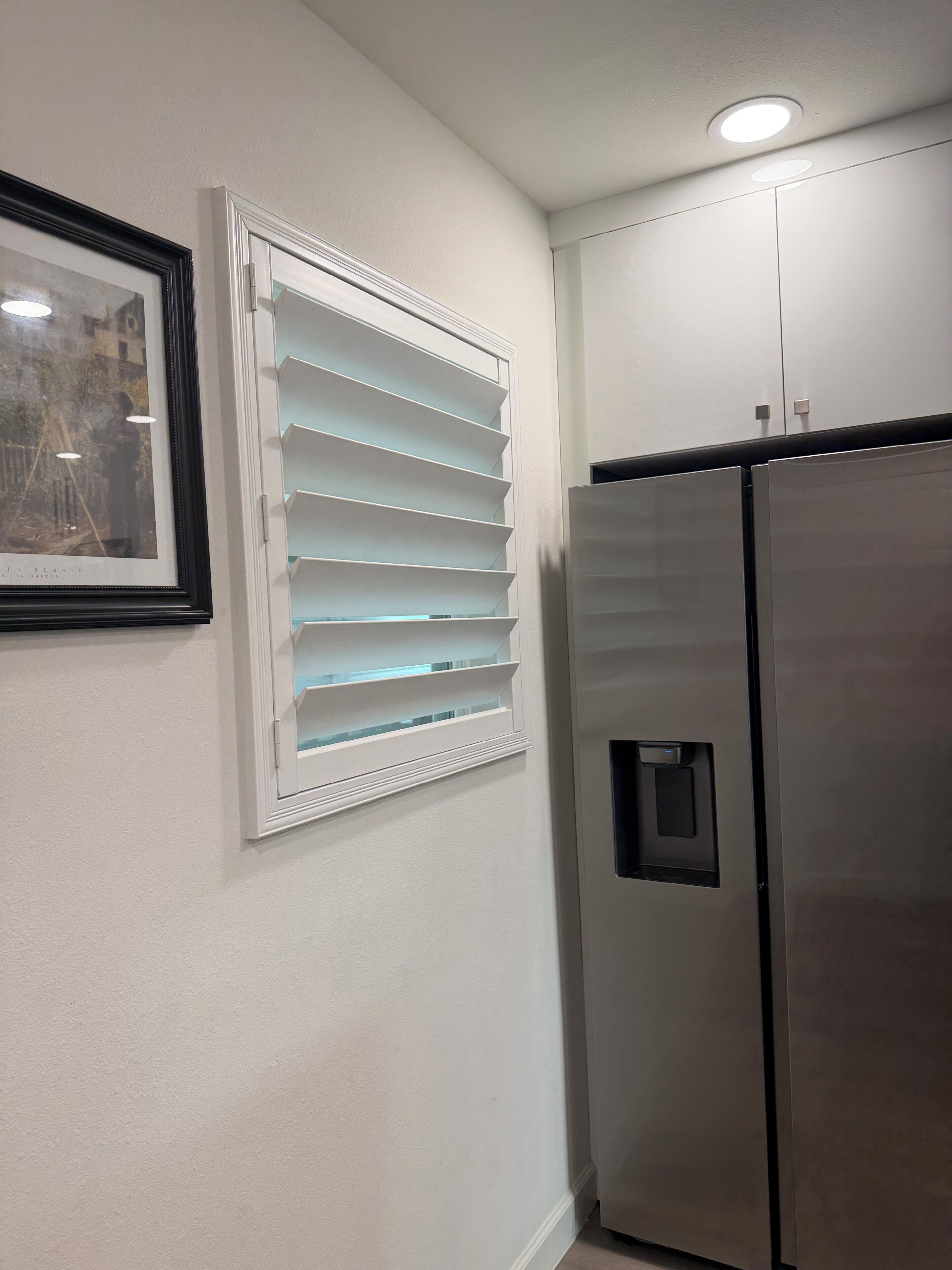 Una cocina con un refrigerador de acero inoxidable, un ventanal blanco con persianas y un cuadro enmarcado.