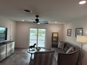 A modern living room with a ceiling fan, a TV, a dining table with a decorative horse figurine, a cushioned seating area, and a decorative stand with flowers.
