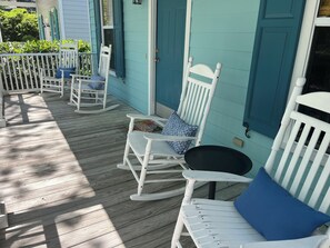 Eine Veranda mit weißen Schaukelstühlen und einem Tisch, einer türkisblauen Tür und einem weißen Geländer.