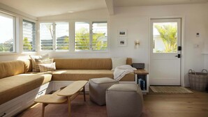 Ein modernes Wohnzimmer mit einem Ecksofa, Poufs, einem kleinen Tisch und einer Tür mit Blick auf Palmen.