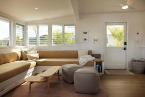 Ein modernes Wohnzimmer mit einem Ecksofa, Poufs, einem kleinen Tisch und einer Tür mit Blick auf Palmen.
