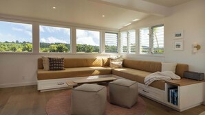 Ein modernes Wohnzimmer mit einer Eckcouch, Hocker, einem Couchtisch und großen Fenstern mit Blick ins Grüne.