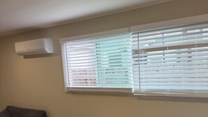 Una habitación con una mesa y sillas de madera, un sofá gris, una ventana con persianas y un equipo de aire acondicionado en la pared.