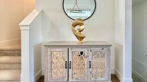 A hallway with a white wall, a wooden staircase, a white console table with a gold abstract sculpture, and a round mirror with a chandelier reflection. - Luxury 3Br, Oasis Gated, Yard Pets Welcome (Langley Township)