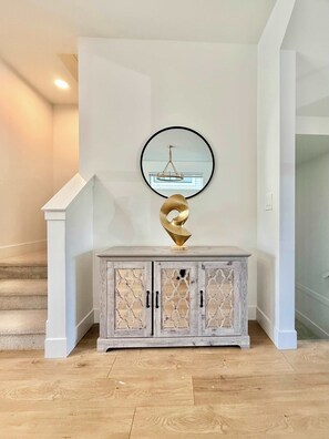 Ein Flur mit weißer Wand, einer Holztreppe, einem weißen Konsolentisch mit einer goldenen abstrakten Skulptur und einem runden Spiegel mit einem Chandelier-Spiegelbild.