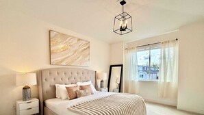 A bedroom with a large bed, a nightstand, a bedside lamp, a wall-mounted light fixture, a window with curtains, and a painting on the wall. - Luxury 3Br, Oasis Gated, Yard Pets Welcome (Langley Township)