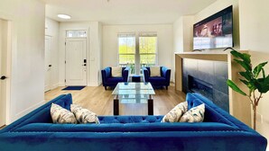 A living room with a blue sofa, a glass coffee table, a fireplace, and a TV mounted on the wall. - Luxury 3Br, Oasis Gated, Yard Pets Welcome (Langley Township)