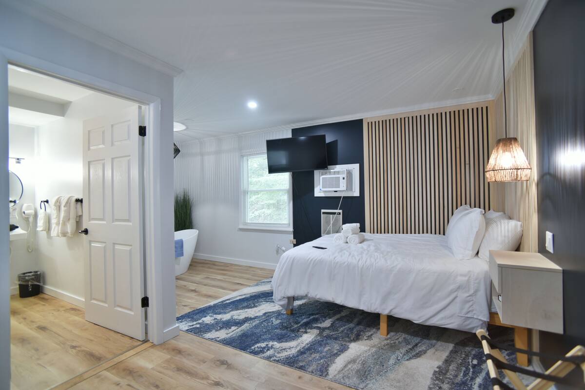 Un dormitorio con cama, mesita de noche, lámpara, televisor, equipo de aire acondicionado y una puerta que da a otra habitación.