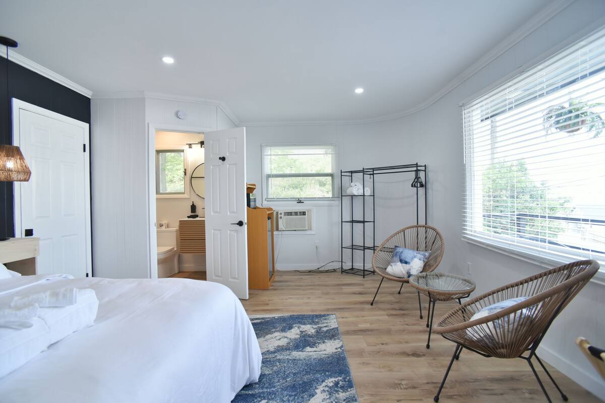 Un dormitorio con una cama, una silla, una mesita, una ventana con persianas y una puerta que da a un baño.