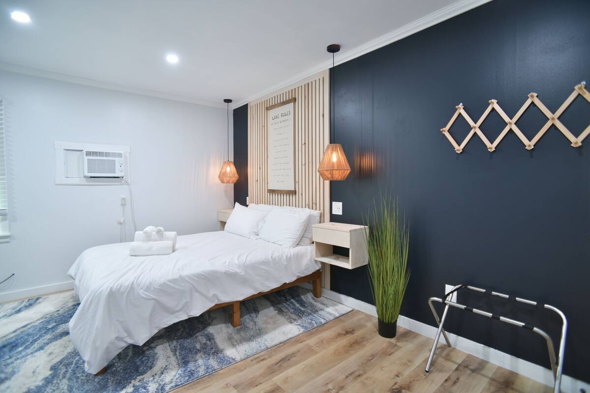 Un dormitorio moderno con cama, mesitas de noche, aire acondicionado de pared y una lámpara mural, además de un elemento decorativo en la pared.