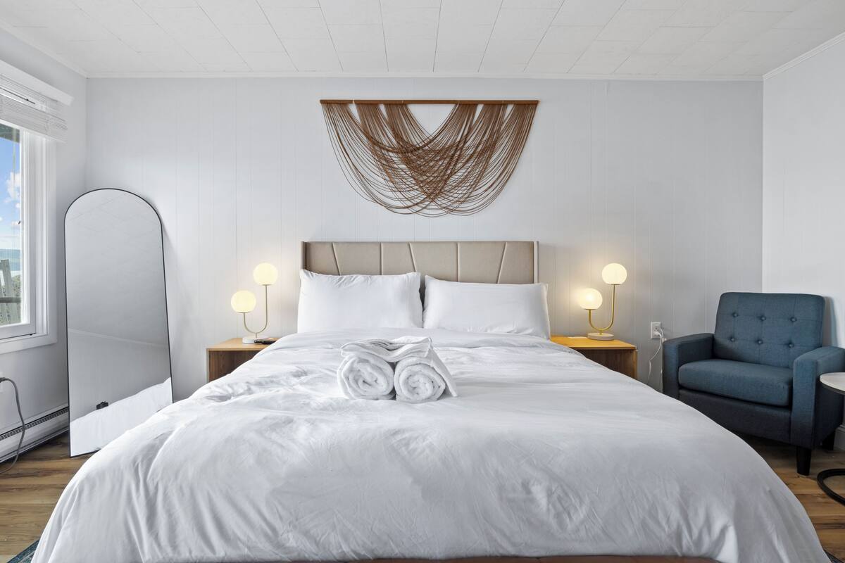 Una cama bien tendida con sábanas blancas, cabecero, lámparas de noche y un sillón azul en una habitación de hotel moderna.