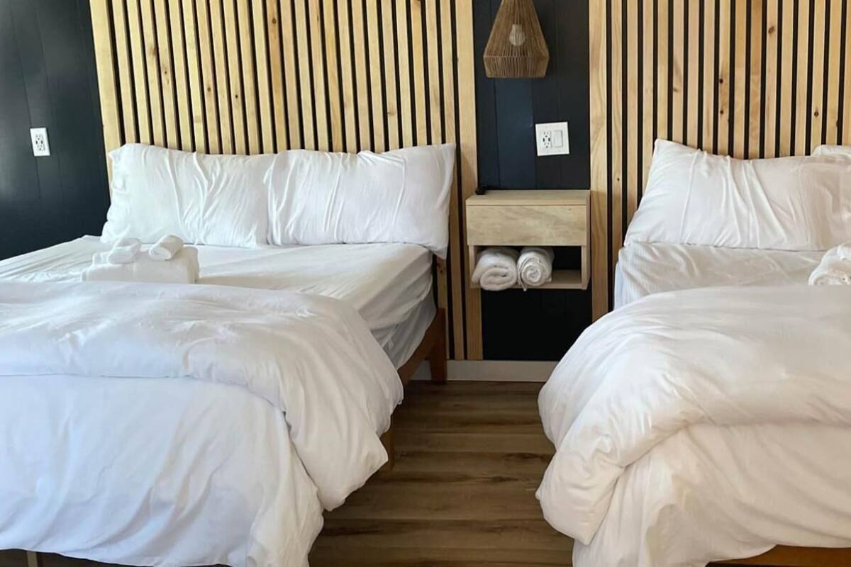 Una habitación con dos camas, cada una con ropa de cama y almohadas blancas, un cabecero de madera y una mesita de noche de madera con toallas.