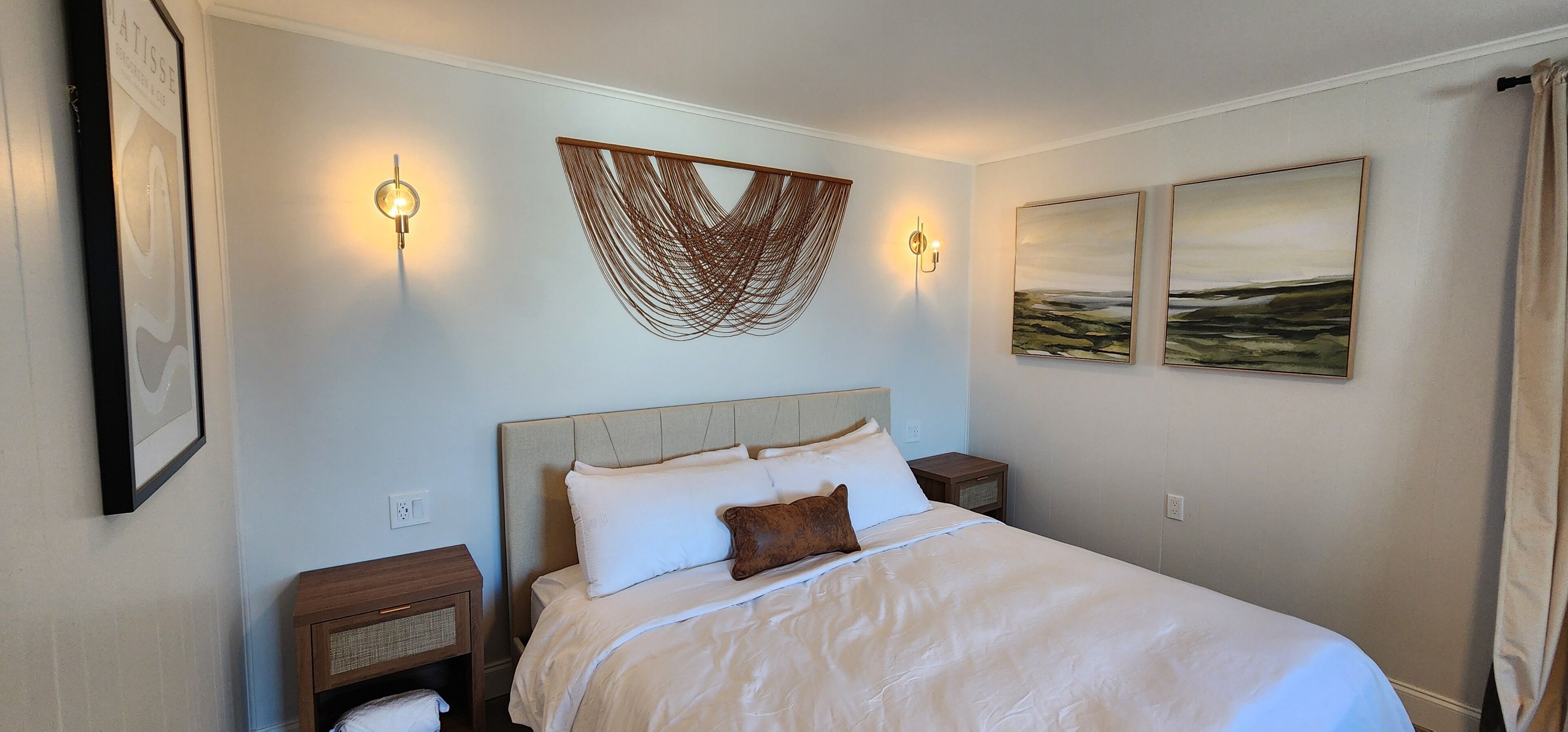 Una cama bien hecha con ropa de cama blanca, dos lámparas fijadas a la pared y dos obras de arte enmarcadas.