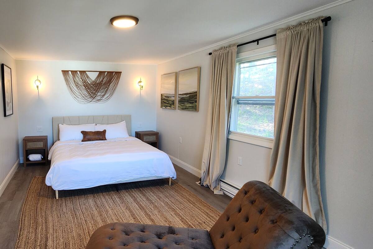 Una habitación de hotel con una cama grande, una zona de estar con una silla, un espejo, una mesita de noche y una ventana con cortinas.