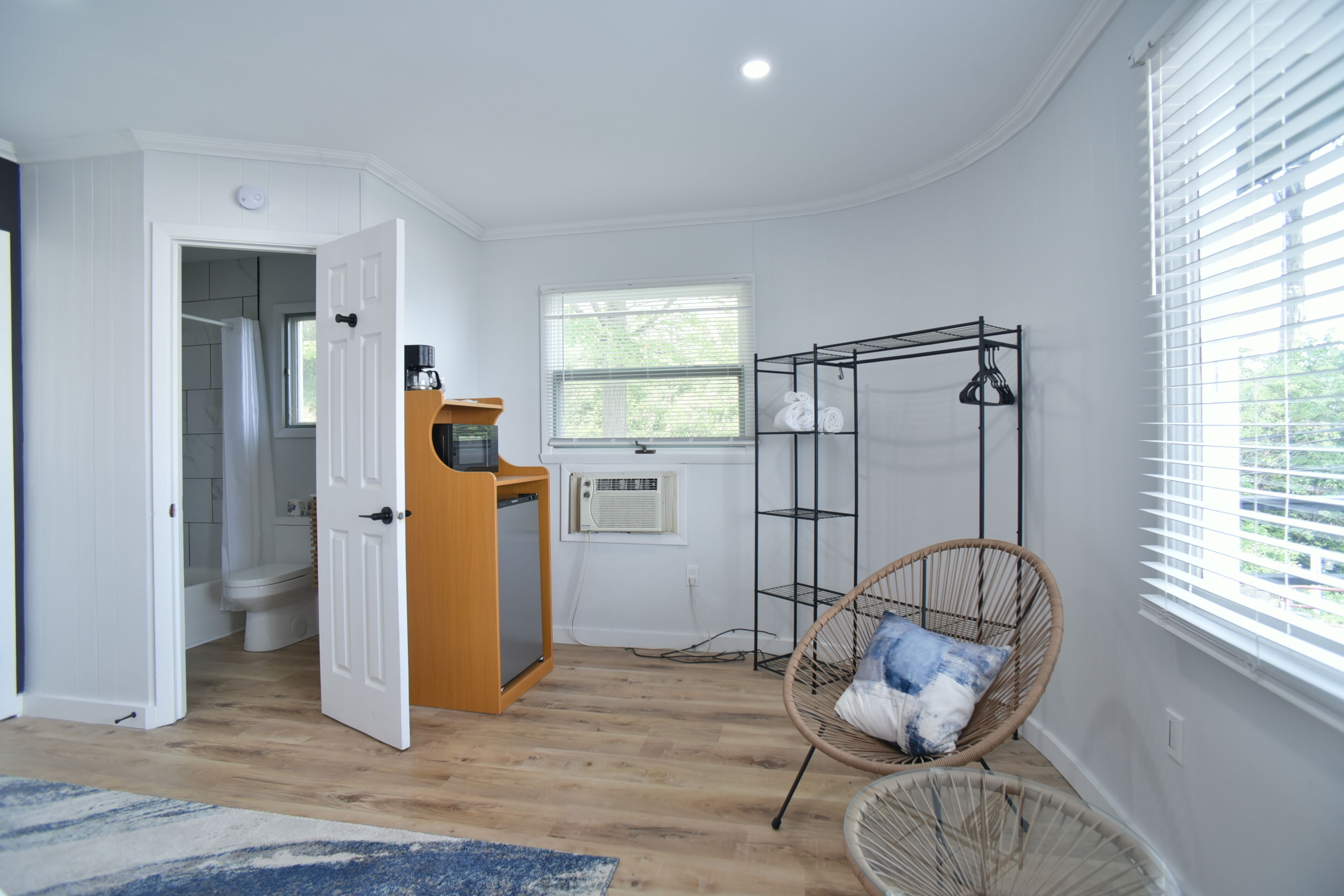 A room with a wooden floor, a white door leading to a bathroom, a radiator, a wicker chair, and a window with blinds.
