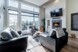 A modern living room with a fireplace, leather sofas, a flat-screen TV, and large windows offering a view of trees and mountains. - Chalet Altitude near Le Massif de Charlevoix (Petite-Rivière-Saint-François)