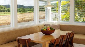 A dining area with a wooden table and chairs, a bowl of fruit, and a view of a field through large windows.