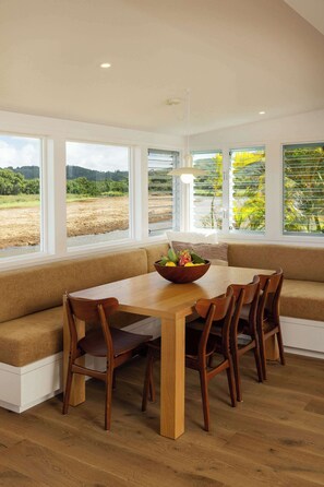 A dining area with a wooden table and chairs, a bowl of fruit, and a view of a field through large windows. - Cottage Four - Luxury Riverfront Stay on Kauai (Hanalei)