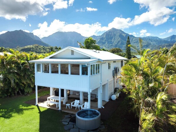 A white two-story house with a covered patio, a hot tub, and a view of mountains. - Cottage Four - Luxury Riverfront Stay on Kauai (Hanalei)