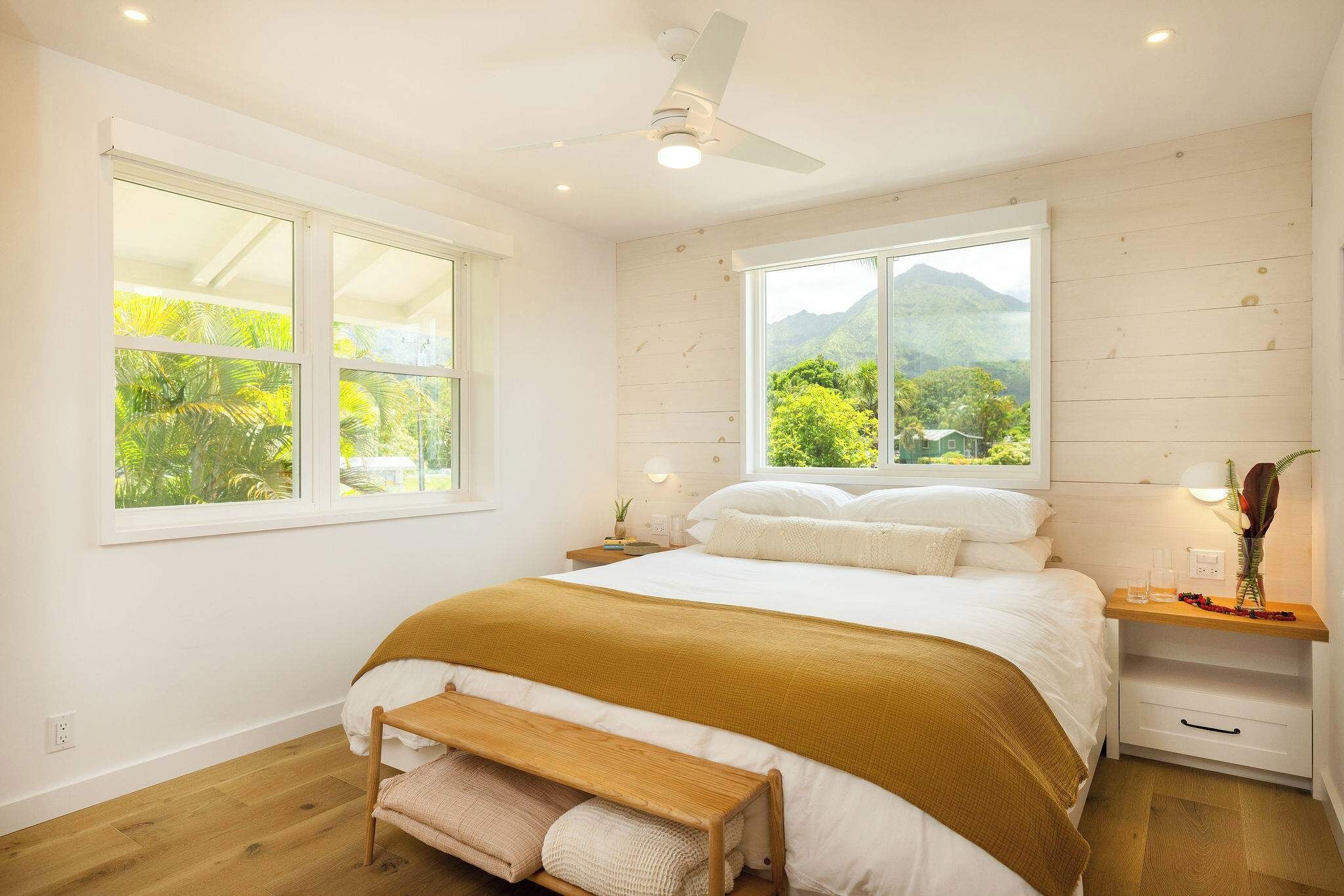 A bedroom with a bed, a nightstand, a bench, a ceiling fan, and large windows with a view of greenery and mountains.