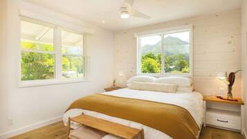 A bedroom with a bed, a nightstand, a bench, a ceiling fan, and large windows with a view of greenery and mountains.
