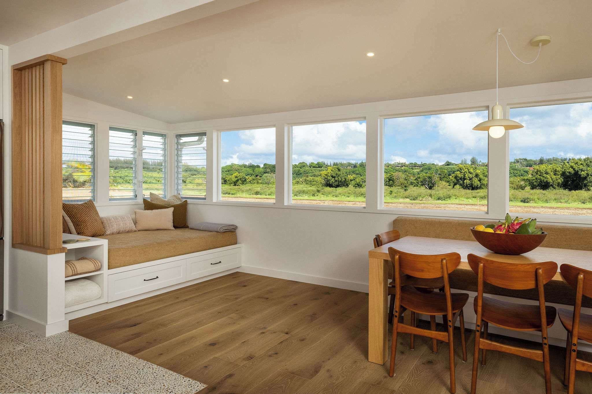 A room with a large window, a wooden bench, a dining table with chairs, and a hanging light fixture.