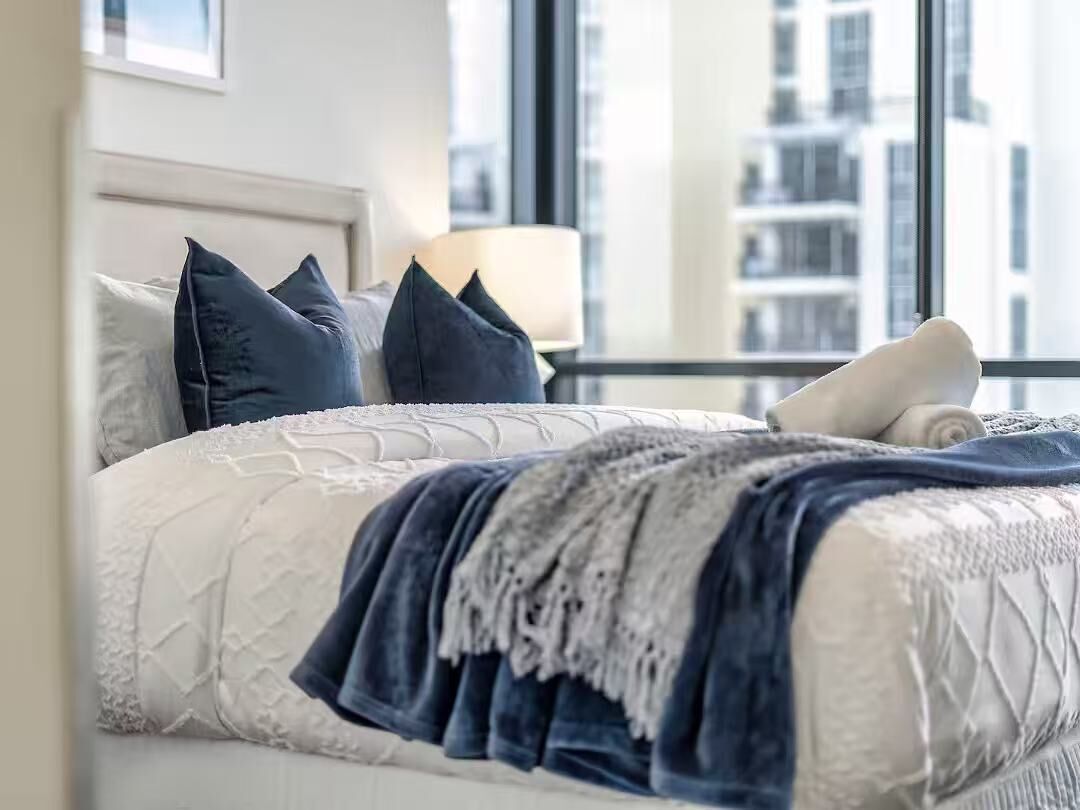 Un lit bien fait, avec une couette blanche et bleue, un plaid gris et des oreillers foncés.