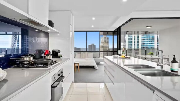 A modern kitchen with a stove, oven, and sink, a dining area with a view of the city, and a large window. - Scenic View 2 Bedroom Apartment - Pools, Gym and Secure Parking (Surfers Paradise)