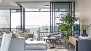 A modern living room with a sofa, a small table, and chairs, large windows, and potted plants. - Scenic View 2 Bedroom Apartment - Pools, Gym and Secure Parking (Surfers Paradise)