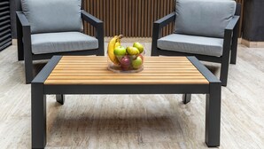 Un espace extérieur moderne aménagé avec deux fauteuils gris à revêtement rembourré, une table en bois et un saladier rempli de bananes, de pommes et de poires.