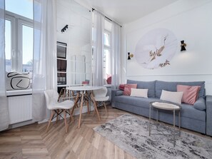 En moderne stue med en grå sofa, hvide stole, et rundt bord og et stort vindue med lette gardiner.