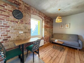 Ein gemütliches Wohnzimmer mit Backsteinwand, einer Couch, einem Esstisch mit Stühlen, einer Uhr und einem Fenster mit Blick.