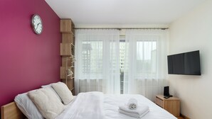 Ein modernes Hotelzimmer mit Bett, Fernseher, einer Uhr und Blick auf Gebäude durch leichte Vorhänge.