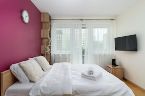 Habitación de hotel moderna con cama, televisor, reloj y vista a edificios a través de cortinas translúcidas.