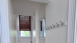 Um corredor com escadas de madeira, uma janela com persianas e um aparador de casacos com ganchos.