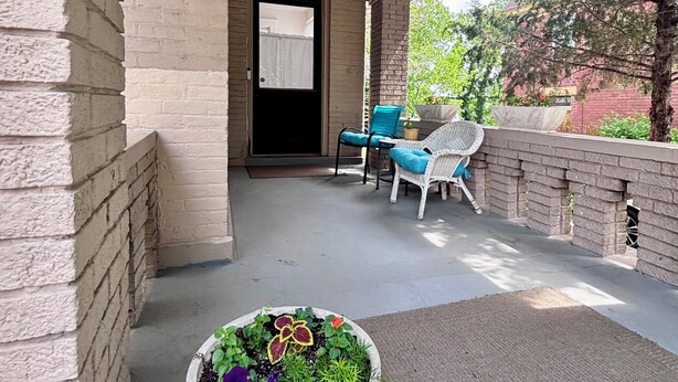 Varanda coberta com piso de concreto, vaso com flores, duas cadeiras de vime e um lustre.